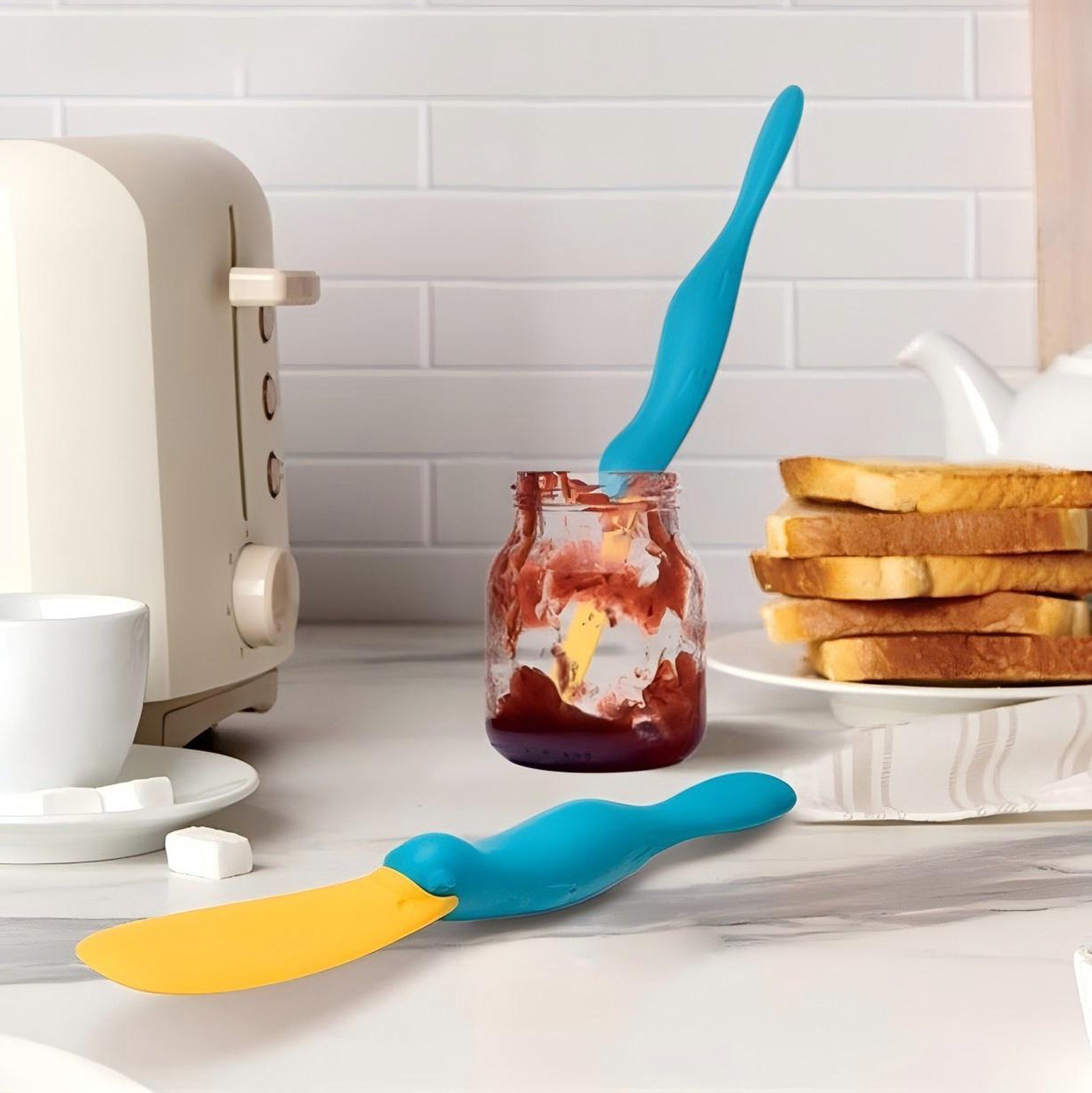 Blue and yellow spatulas on a kitchen counter with toast and jam. platypus jar scraper, peanut butter scraper tool, waste free kitchen gadget, OTOTO jar spatula, jam spreader Pakistan